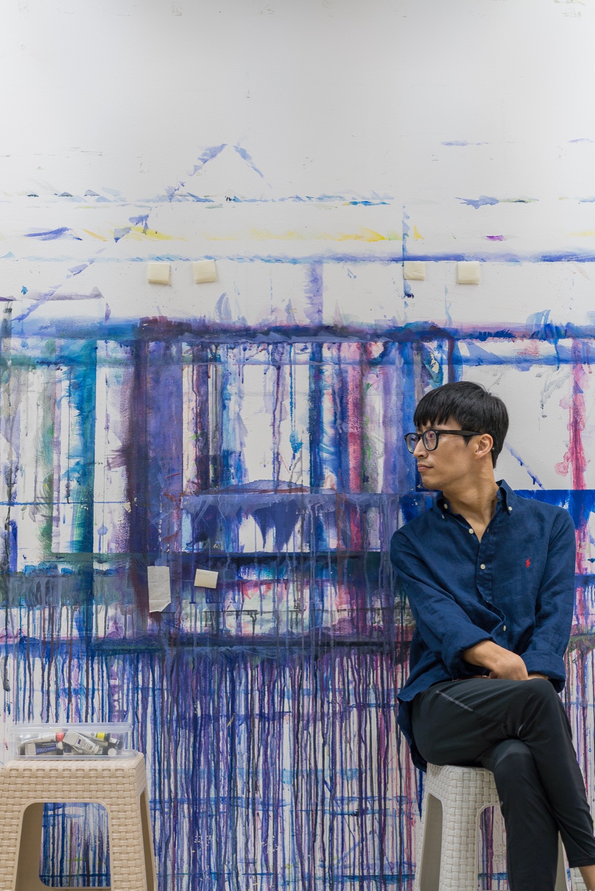 The artist sitting on a stool in front of the studio wall, looking off to one side. Behind him, paint has run down the wall in blue and violet streaks.